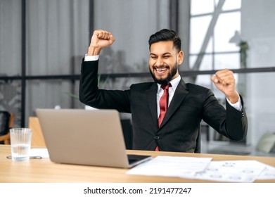 Excited indian or arabian businessman in suit, sits in a modern office, looking at laptop, happy by good news, rejoice by win, raising hand in yes gesture, celebrating project success, big profit - Powered by Shutterstock