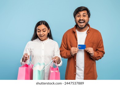 Excited Indian couple with colorful shopping bags and a blue credit card, portraying consumerism on blue - Powered by Shutterstock