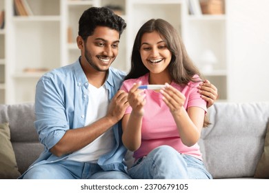 Excited indian couple looking at positive pregnancy test, smiling and enjoying great news, sitting on couch at home. Loving hindu spouses feeling happy about future parenthood - Powered by Shutterstock