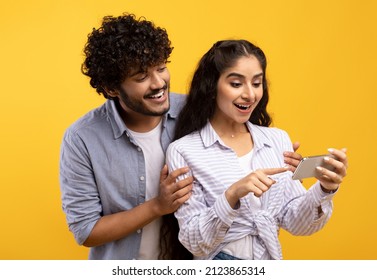 Excited Indian Couple Looking At Smartphone Screen, Using New App Or Reading Cool News, Standing Over Yellow Studio Background. Happy Man And Woman Using Cellphone, Guy Embracing Girlfriend