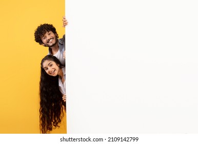 Excited indian couple standing behind white advertisement board and peeking out, showing empty space for your design or offer, posing over yellow studio background, free space