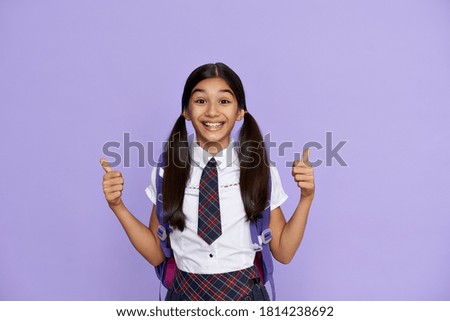 Excited indian kid primary school girl with backpack wearing uniform showing thumbs up isolated on violet background. Happy latin child student celebrating freedom, recommending best education choice.