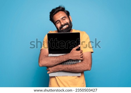 Excited indian man hugging laptop with black blank screen, holding it tight near chest and smiling, male technology lover posing isolated over blue studio background, copy space