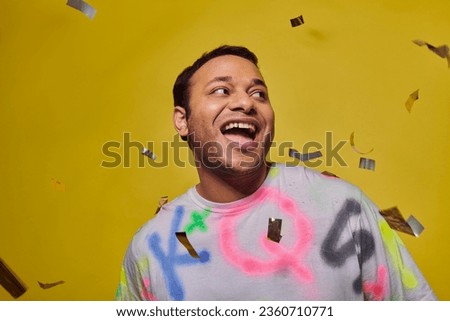 excited indian man smiling near falling confetti on yellow backdrop, party concept, happy face
