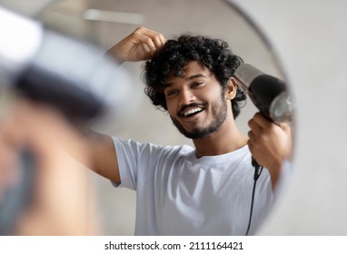 Excited indian man using hairdryer after shower, drying his curly hair, making hairdo, taking care of himself in the morning, smiling at his reflection in mirror. Everyday hygiene concept