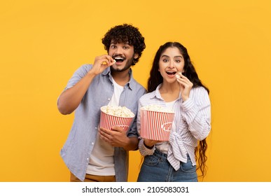 Excited indian man and woman eating popcorn at cinema, standing over yellow studio background. Emotional couple watching comedy movie at cinema or at home, entertainment concept