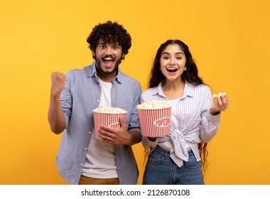 Excited indian man and woman eating popcorn, watching movie or football game, celebrating success of their team over yellow studio background