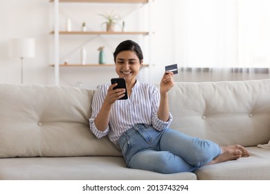 Excited Indian Woman Paying Online By Credit Card For Goods, Using Smartphone, Sitting On Couch At Home, Happy Young Female Satisfied Customer Browsing Internet Banking Service On Device, Shopping