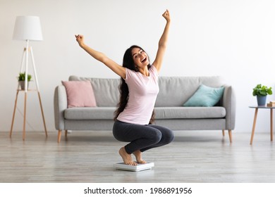 Excited Indian woman sitting on scales, raising hands up, happy with result of her weight loss program at home. Young Asian lady overjoyed over achieving her slimming goal, full length