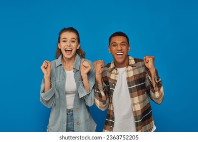 excited interracial couple screaming from joy looking at camera on blue backdrop, emotional reaction