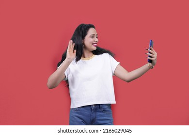 Excited Joyful Beautiful Indian Girl Or Young South Asian Woman Wearing White T Shirt Standing Doing Selfie Shot On Mobile Phone. Woman Talking In Video Call Waving Hands..