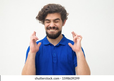 Excited Joyful Guy Keeping Fingers Crossed, Making Wish. Handsome Bearded Young Man In Blue Casual T-shirt Posing Isolated Over White Background. Eager Concept