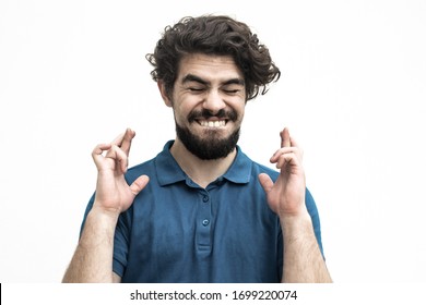 Excited Joyful Guy Keeping Fingers Crossed, Making Wish. Handsome Bearded Young Man In Blue Casual T-shirt Posing Isolated Over White Background. Eager Concept