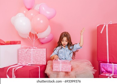 Excited Joyful Little Girl In Tulle Skirt Opening Birthday Present Surround Big Colorful Gift Boxes Isolated On Pink Background. Celebrating Birthday Party With Balloons And Gifts Of Amazing Kid