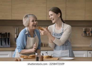 Excited joyful senior mother and happy adult daughter woman preparing sandwiches in home kitchen, having fun, laughing, tasting meal, giving snack to bite, enjoying culinary hobby - Powered by Shutterstock