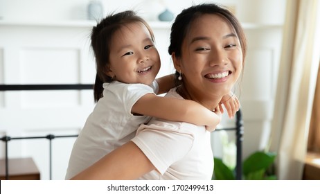 Excited kid girl play with Asian mother at home. Cheery mom piggybacks cute child carrying little daughter on back having fun on weekend spending time together. Happy family close up portrait concept - Powered by Shutterstock