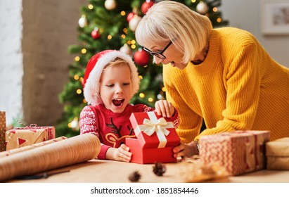 Excited Kid In Santa Hat Opening Gift Box From Granny While Celebrating Christmas Holiday At Home
