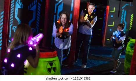 Excited kids and theirs parents aiming laser guns at other players during lasertag game in dark room