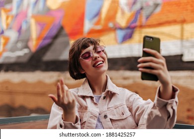 Excited lady with short hair taking photo outdoors. Brunette teen girl in light jacket and pink glasses making selfie and smiling in city.. - Powered by Shutterstock - Get 15% off with code: PIKWIZARD15