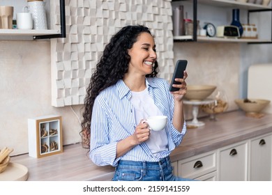 Excited latin woman using smartphone while drinking morning tea in kitchen at home, positive lady reading text messages on her cellphone and smiling - Powered by Shutterstock