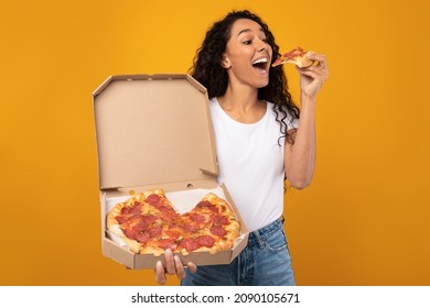 Excited Latin Young Lady Enjoying Pizza Holding And Biting Tasty Slice Posing With Carton Box Over Yellow Orange Studio Background. Junk Food Lover Eating Italian Pizza. Unhealthy Nutrition Cheat Meal - Powered by Shutterstock