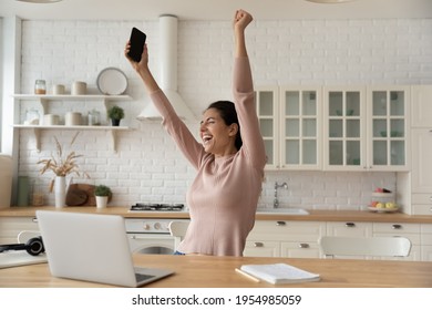 Excited Latina Woman Freelancer Raise Hands Up Hold Phone Scream Hooray In Delight Celebrate Finish Work On Difficult Project. Euphoric Young Lady Remote Employee Meet Deadline With All The Work Done