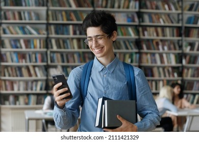 Excited Laughing Gen Z Smart Young Student Boy In Glasses Holding Stacked Books In Library, Using Online App For Study On Mobile Phone, Reading Text Message, Making Video Call, Watching Content