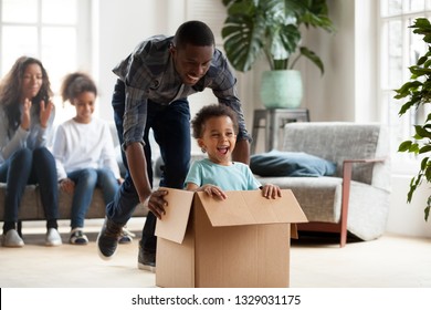 Excited Little Black Boy Sit In Cardboard Box Playing With Young Dad Unpacking Packages On Moving Day, Happy Young African American Family Have Fun Entertain With Small Kids Relocating To New Home