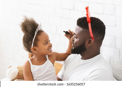 Excited little black girl putting make up on her father face, both wearing plastic crowns, close up