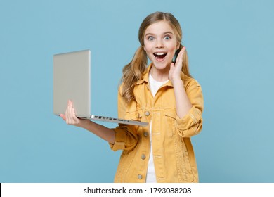 Excited Little Blonde Kid Girl 12-13 Years Old In Yellow Jacket Isolated On Pastel Blue Wall Background. Childhood Lifestyle Concept. Mock Up Copy Space. Working On Laptop Pc Computer Spreading Hands