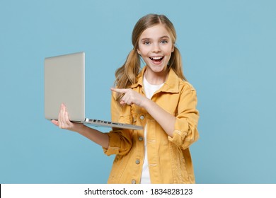 Excited little blonde kid girl 12-13 years old in yellow jacket isolated on pastel blue wall background. Childhood lifestyle concept. Mock up copy space. Pointing index finger on laptop pc computer