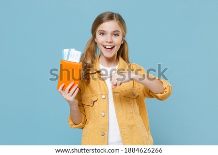Excited little blonde kid girl 12-13 years old isolated on blue background. Passenger traveling abroad to travel on weekends getaway. Air flight journey concept. Point index finger on passport ticket