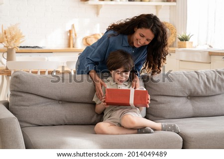 Excited little boy receiving birthday present from mum sitting on sofa in living room. Children holiday celebration concept with loving mother giving gift to adorable preschool son congratulating kid