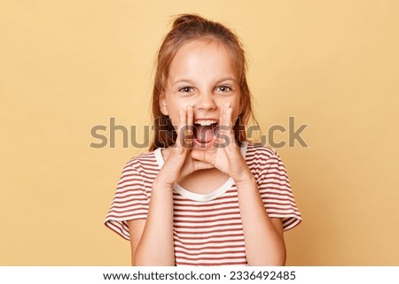 Excited little brown haired girl wearing striped t-shirt standing isolated over beige background keeps hands near mouth screaming loud important information.
