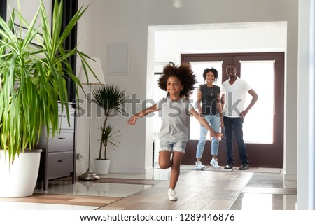 Excited little funny african girl running exploring big modern house moving in, happy black parents and kid daughter coming into new home, cute mixed race child having fun in hallway, family mortgage