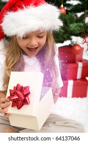 Excited Little Girl Opening Christmas Present With Magical Glow In Front Of The Fir Tree - Closeup