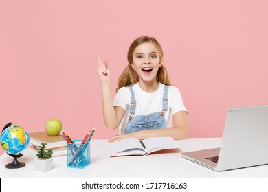 Excited little kid schoolgirl 12-13 years old sit study at white desk with pc laptop isolated on pink background. School distance education at home during quarantine concept. Pointing index finger up