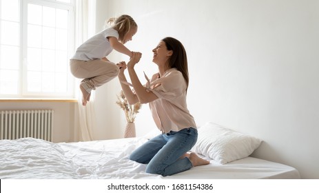 Excited little preschooler girl jump high on comfortable mattress have fun with happy young mom at home, overjoyed small daughter and mother playing engaged in funny activity in bedroom together
