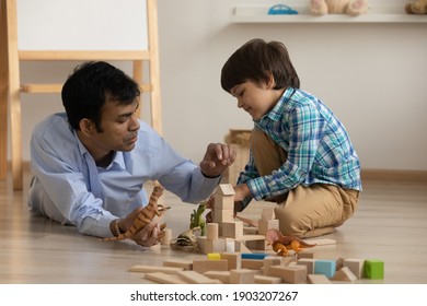 Excited little son and loving dad of indian ethnicity have fun play toys on warm floor at living room. Young male babysitter and preschooler boy engaged in education game with wooden blocks dinosaurs