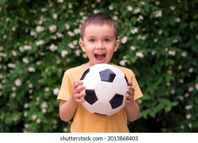 Excited Little Toddler Boy Holding Football Outdoors: Green Leaves And Flowers Behind. The Kid Just Caught Ball And Shouting Of Happiness And Emotions. Active Childhood And Sports Passion Concept