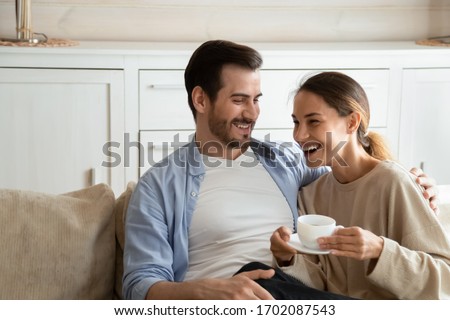 Excited loving family couple resting on cozy sofa with cups of tea coffee, having fun joking in morning. Happy spouses talking chatting laughing, enjoying weekend holiday time together at home.
