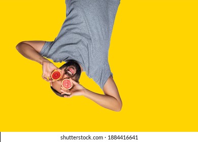 Excited Mad Man Spectator Hanging Upside Down And Looking Through Binoculars From Grapefruit Over Isolated Yellow Studio Background. Watching, Observation, Expression. Copy Space For Text.  
    