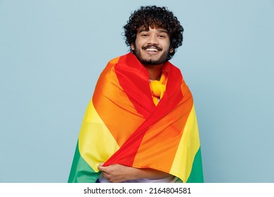 Excited magnificent young bearded Indian man 20s years old wears white t-shirt wrapped in striped colorful rainbow flag looking camera isolated on plain pastel light blue background studio portrait
