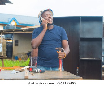 An excited male African Nigerian man or carpenter with a hat, happily holding an hammer and making a call while standing at a business carpentry workshop in readiness to satisfy potential customers - Powered by Shutterstock