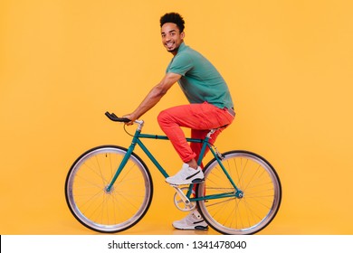 Excited Male Model In Sport White Shoes Posing On Bike. Indoor Photo Of Cheerful African Guy Sitting On Bicycle And Looking To Camera.