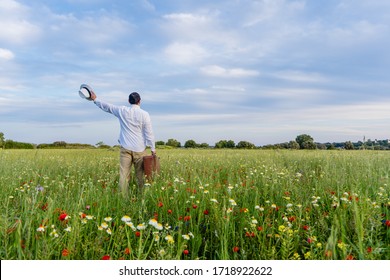 Excited Male On Farmland Sunset Natural Outdoors Background. Unrecognized Hopeful Person Holding Suitcase Backview, Traveling Away Coming Back Scene. White Clouds Blooming Flowers Relaxing Freedom. 