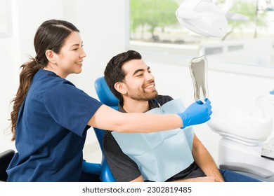 Excited male patient looking at his teeth in the mirror and smiling after doing a whitening or dental treatment at the dentist - Powered by Shutterstock