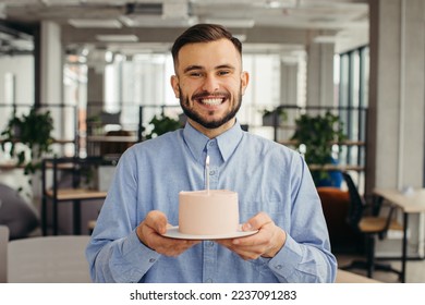 Excited man celebrating birthday in office, looking happy, holding cake with candle, making a wish. - Powered by Shutterstock