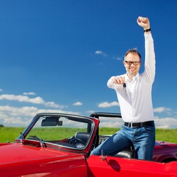 Excited Man Celebrating Ownership Of A Cabriolet Standing With The Hood Down And Door Open Raising His Arm In Jubilation And Holding Out The Key In Sunny Fresh Countryside