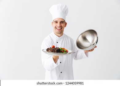 Excited man chef cook wearing uniform opening cloche cover isolated over white background, showing meat dish